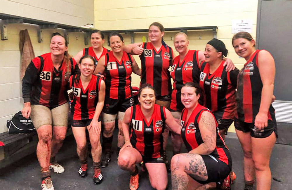 Ten 2019 Blackburn women's development team embracing in red & black uniform in a change room, muddy legs, big grins looking different ways.