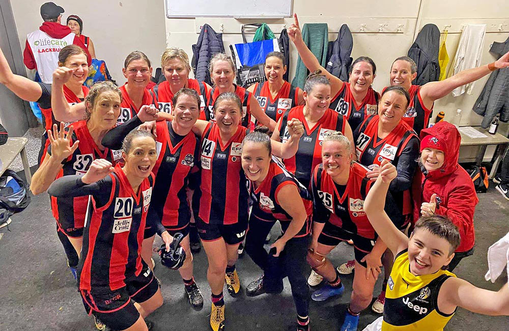 Sixteen 2021 Blackburn Masters women in football change room, jubilant with arms up, smiling. Trainer assisting a girl. A son joins in jubilantly.